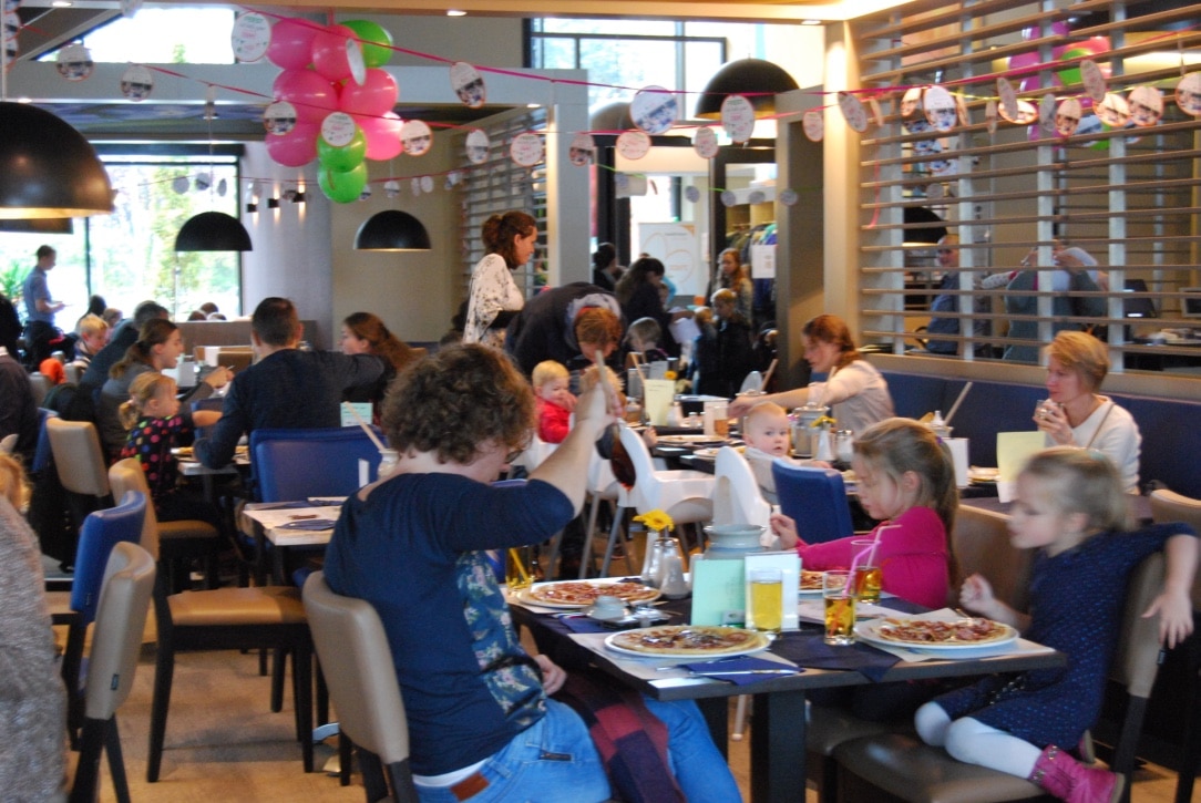 Busy family-friendly restaurant filled with adults and children eating at tables, colorful balloons and party decorations overhead.